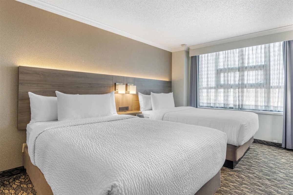 Una habitación de hotel con dos camas, cabecera con iluminación integrada, un ventanal con cortinas y piso alfombrado.