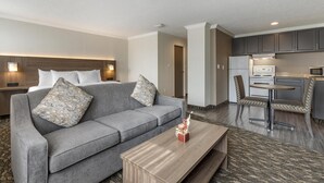 Quarto de hotel com um sofá cinzento, uma mesa de centro de madeira, uma pequena área de cozinha com frigorífico, micro-ondas e uma mesa de jantar com cadeiras.