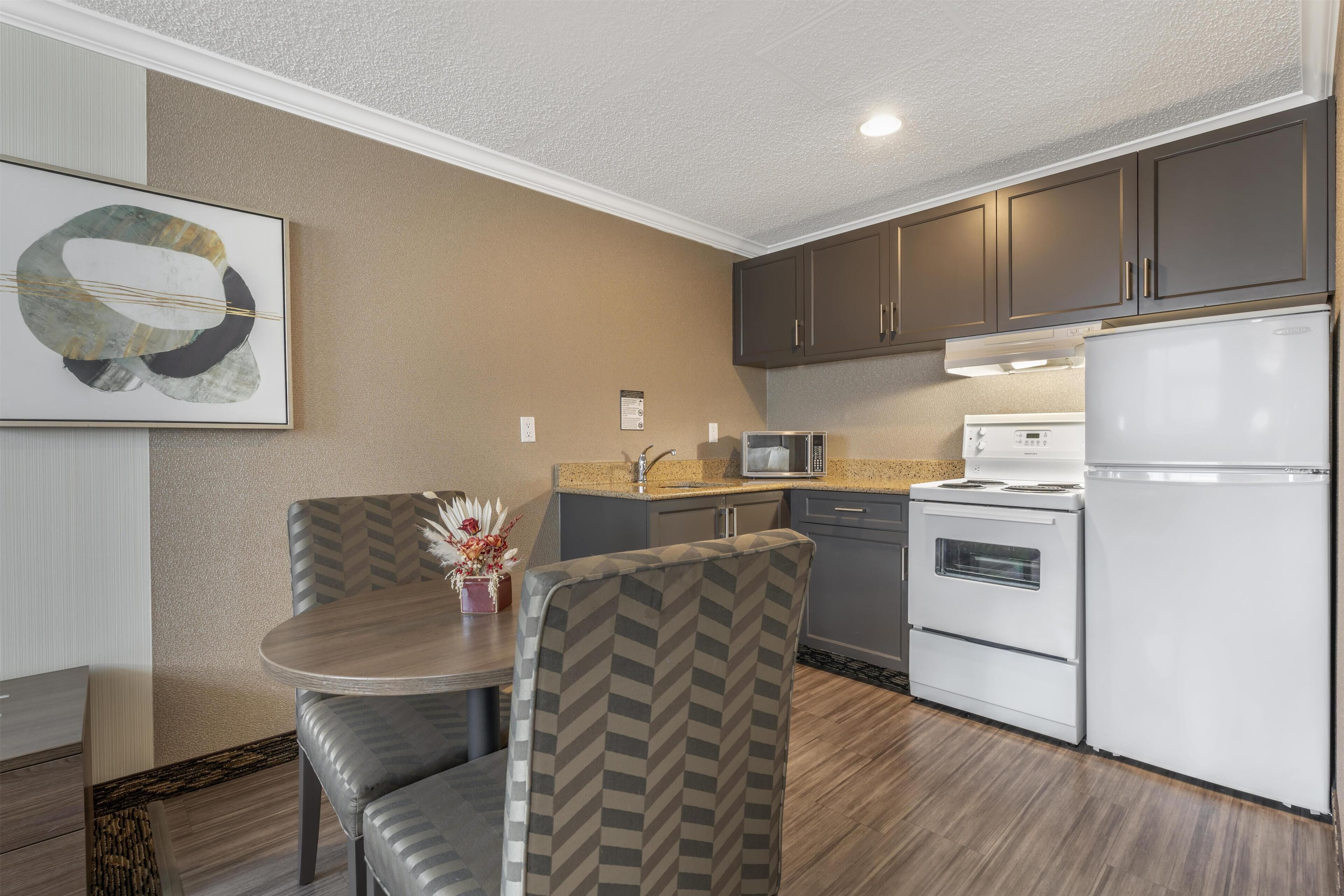 A modern kitchen with dark cabinets, a white refrigerator, a microwave, and a stove. There is a dining area with a table and chairs.