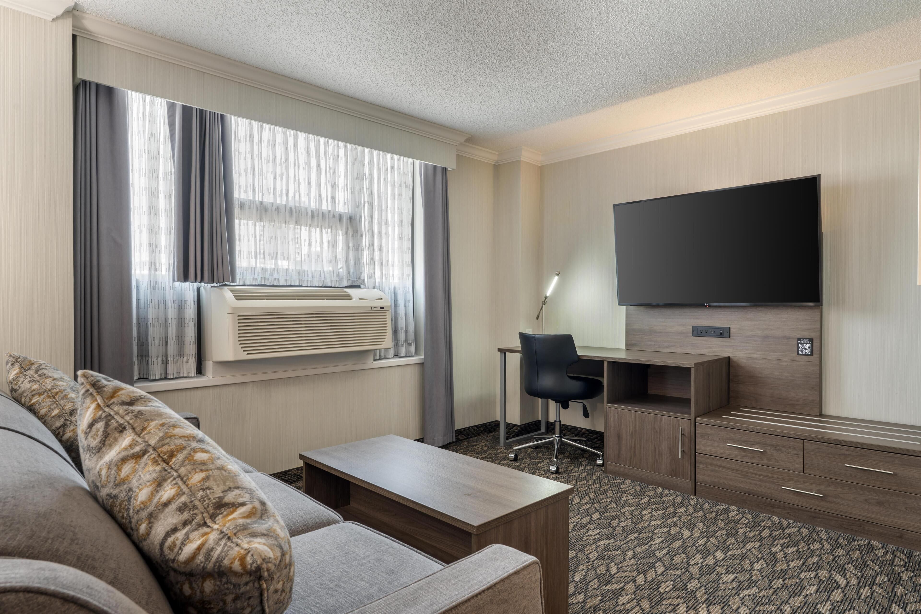 A hotel room with a sofa, a desk, a chair, a flat-screen TV, and a large window with curtains.