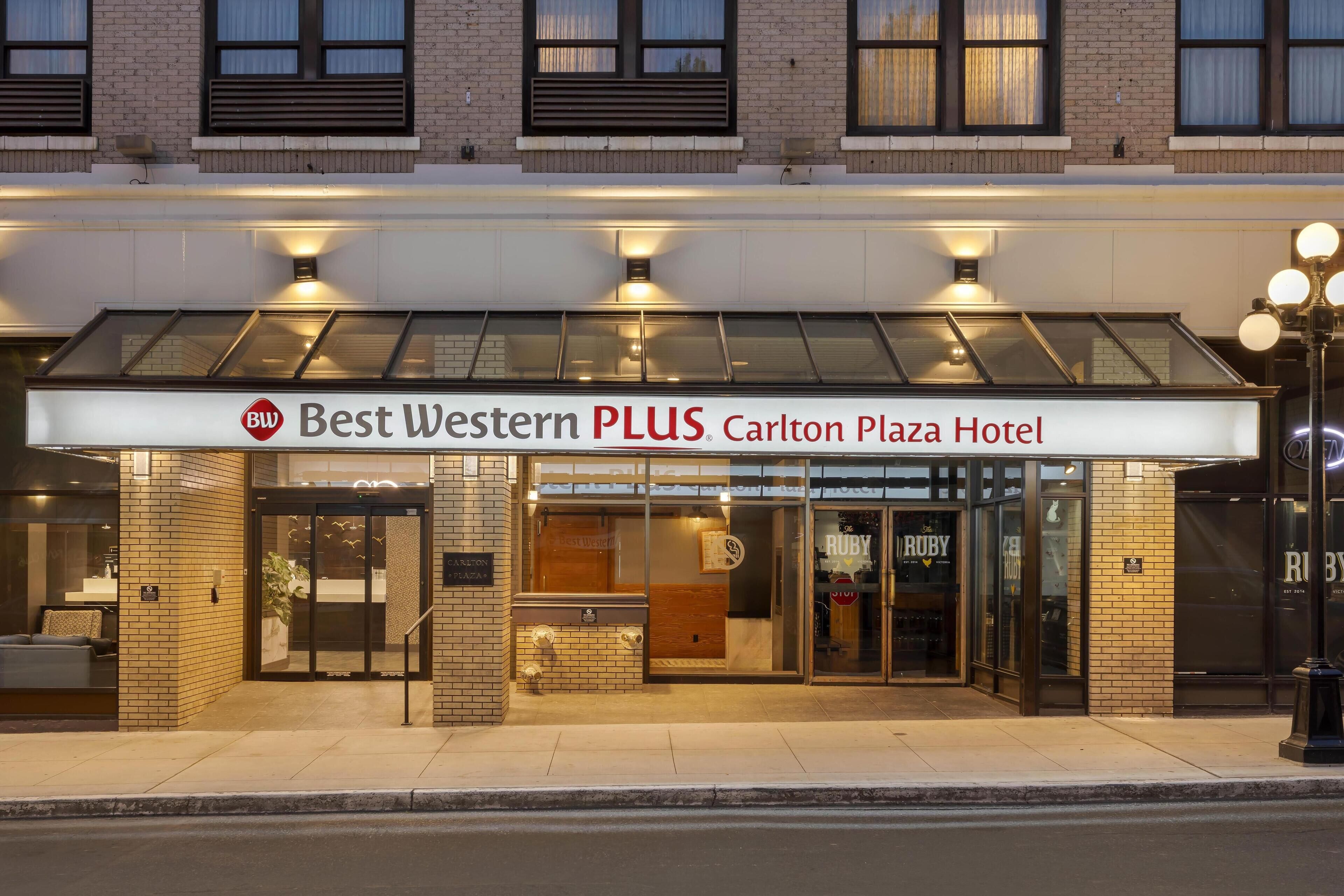 The exterior of the Best Western PLUS Carlton Plaza Hotel with a glass entrance, a signboard, and a brick facade.
