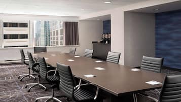 Un salón de conferencias con una larga mesa, numerosas sillas, una estación de café y vistas de edificios altos a través de grandes ventanales.