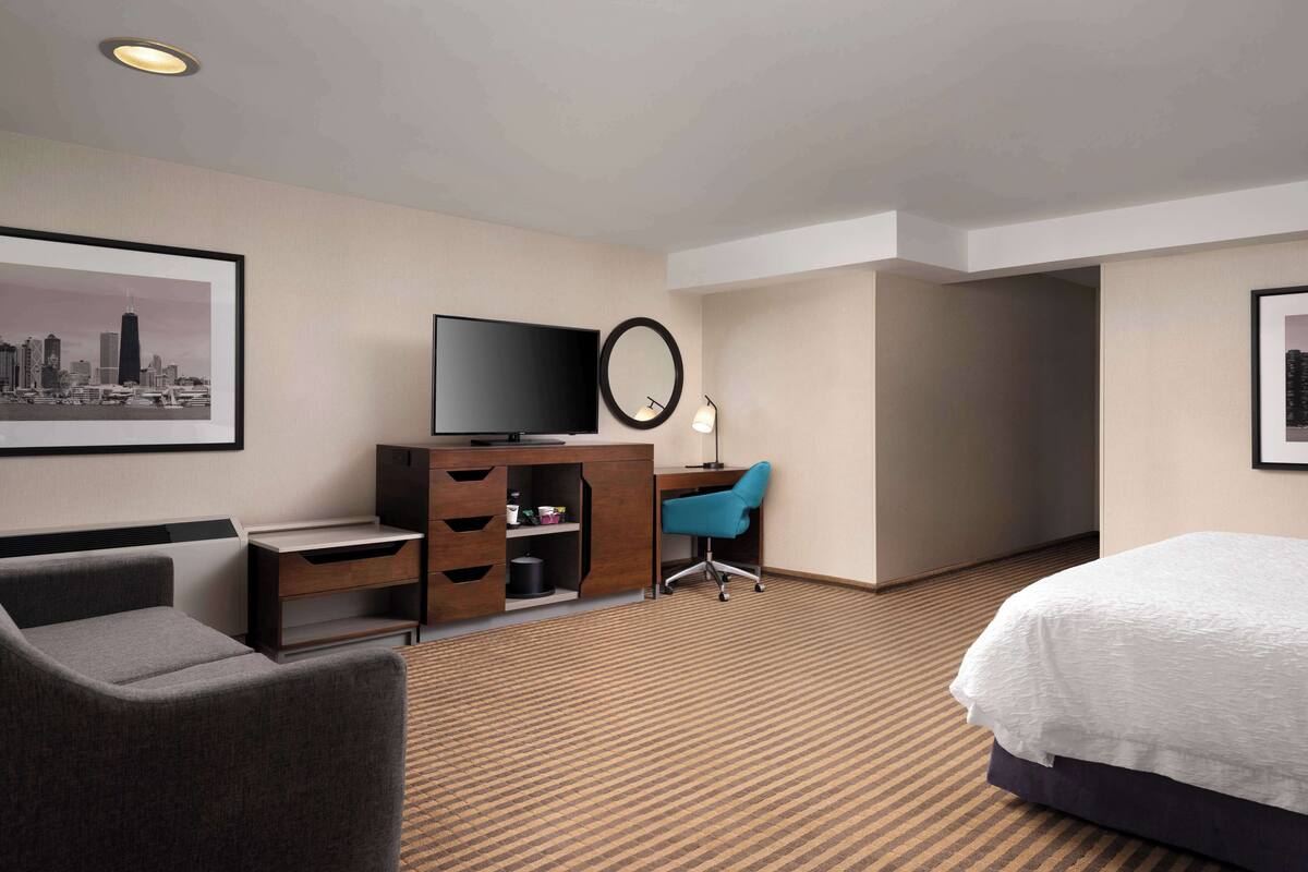 A hotel room with a bed, a desk with a computer, a chair, a TV, and a framed picture on the wall.