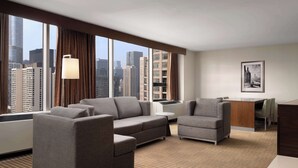 Quarto de hotel com uma grande janela que oferece vista para a cidade, um sofá, uma poltrona, uma mesinha e uma escrivaninha com cadeira.