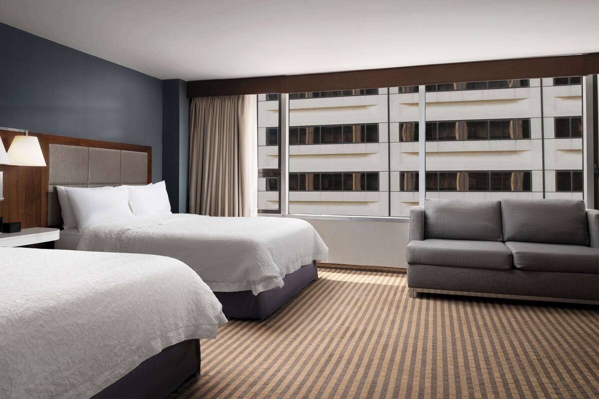 A hotel room with two beds, a sofa, a large window with curtains, and a view of a building.