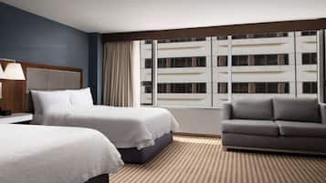 Habitación de hotel con dos camas, un sofá, un ventanal con cortinas y vistas a un edificio.