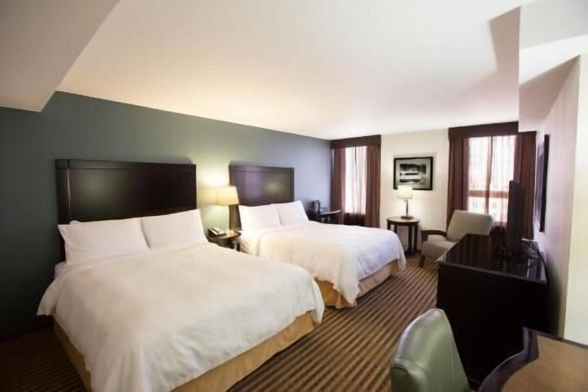 A hotel room with two beds, a desk, a chair, and a lamp.
