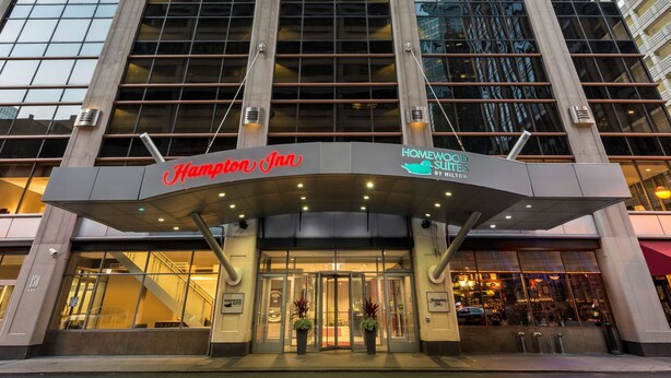 Um hotel com fachada moderna em vidro, com destaque para a placa Hampton Inn e a placa Homewood Suites by Hilton.