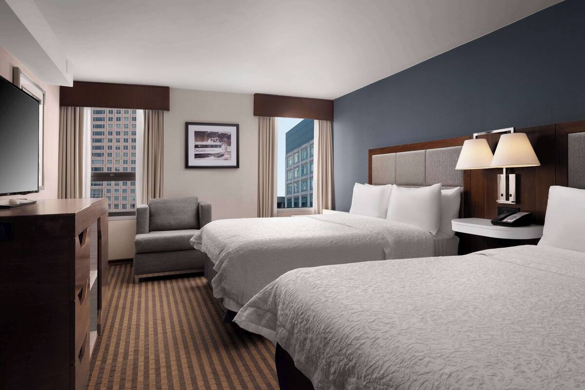 A hotel room with two beds, a desk, a chair, a TV, and a window with curtains.