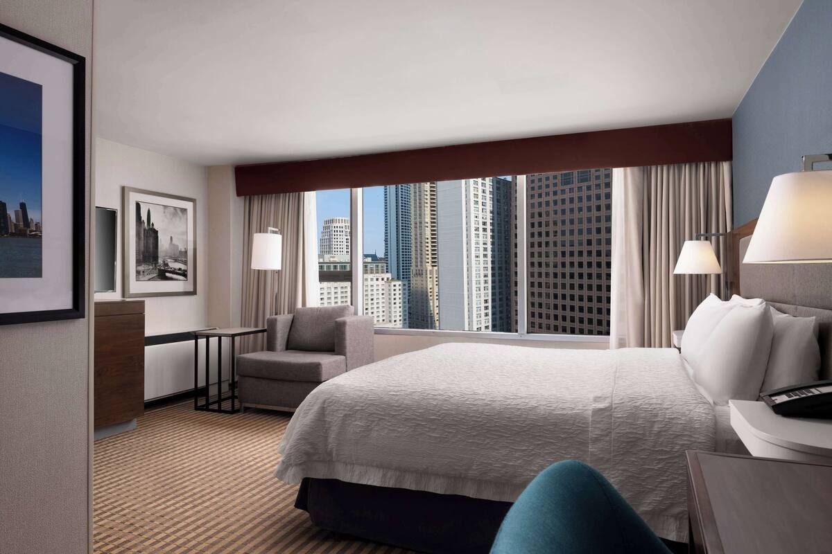 A hotel room with a large bed, a desk, a chair, and a view of tall buildings.
