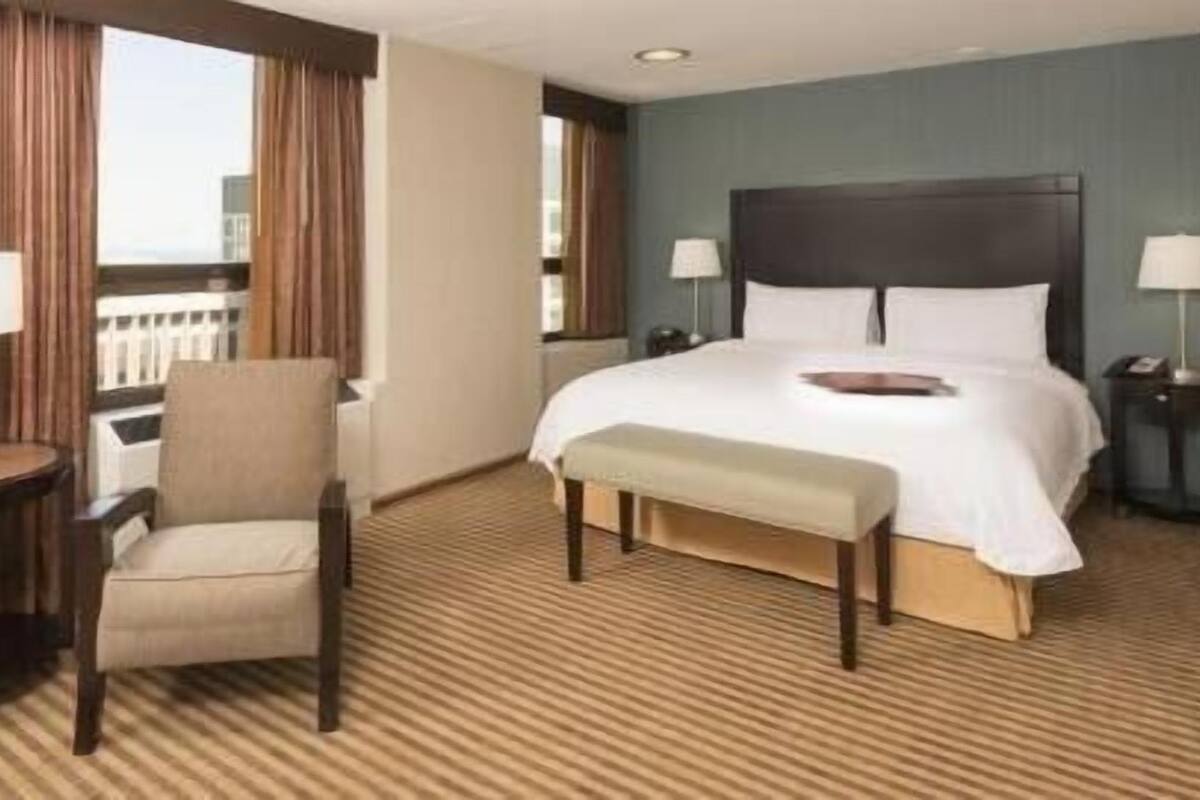 A hotel room with a bed, a chair, a small table, and a window with curtains.