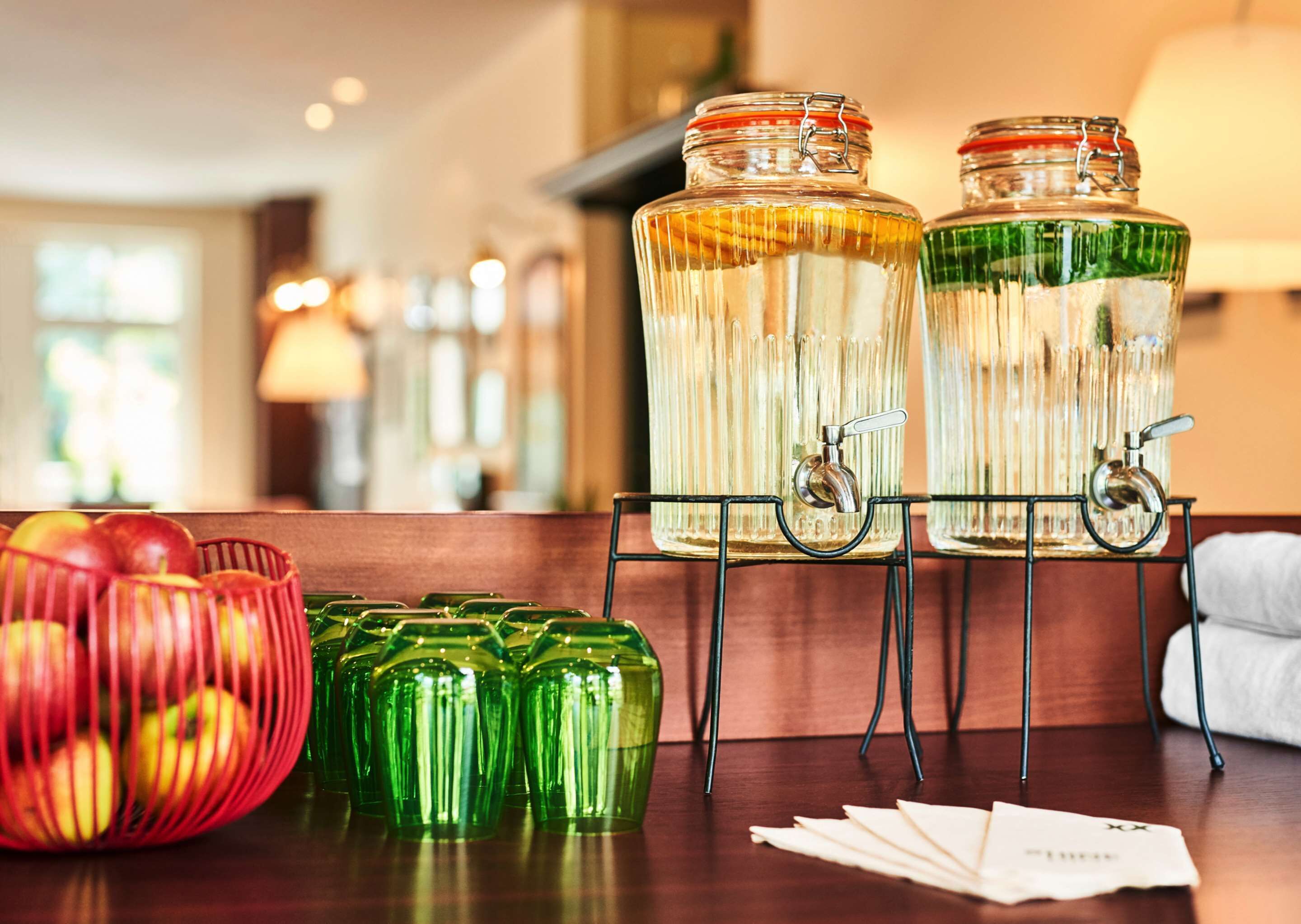 A buffet setup with two glass jars containing food items, a bowl of apples, and a tray of napkins.