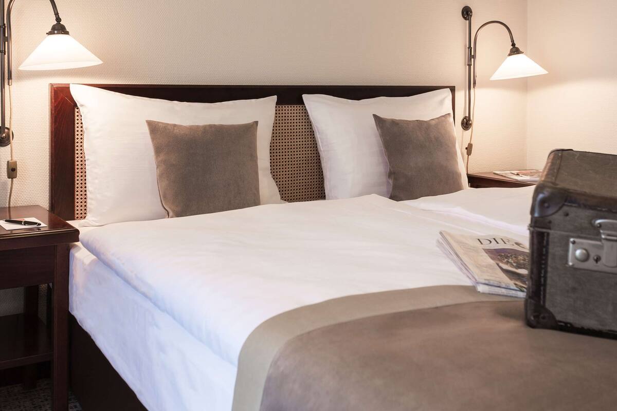 A hotel room with a neatly made bed, two bedside lamps, a nightstand, and a suitcase.