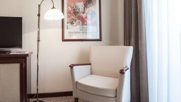 A hotel room with a television on a wooden stand, a floor lamp, a framed movie poster, a beige armchair, and a window with curtains.