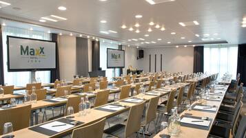 A Maxx Hotel Jena conference room with multiple tables, chairs, and presentation screens.