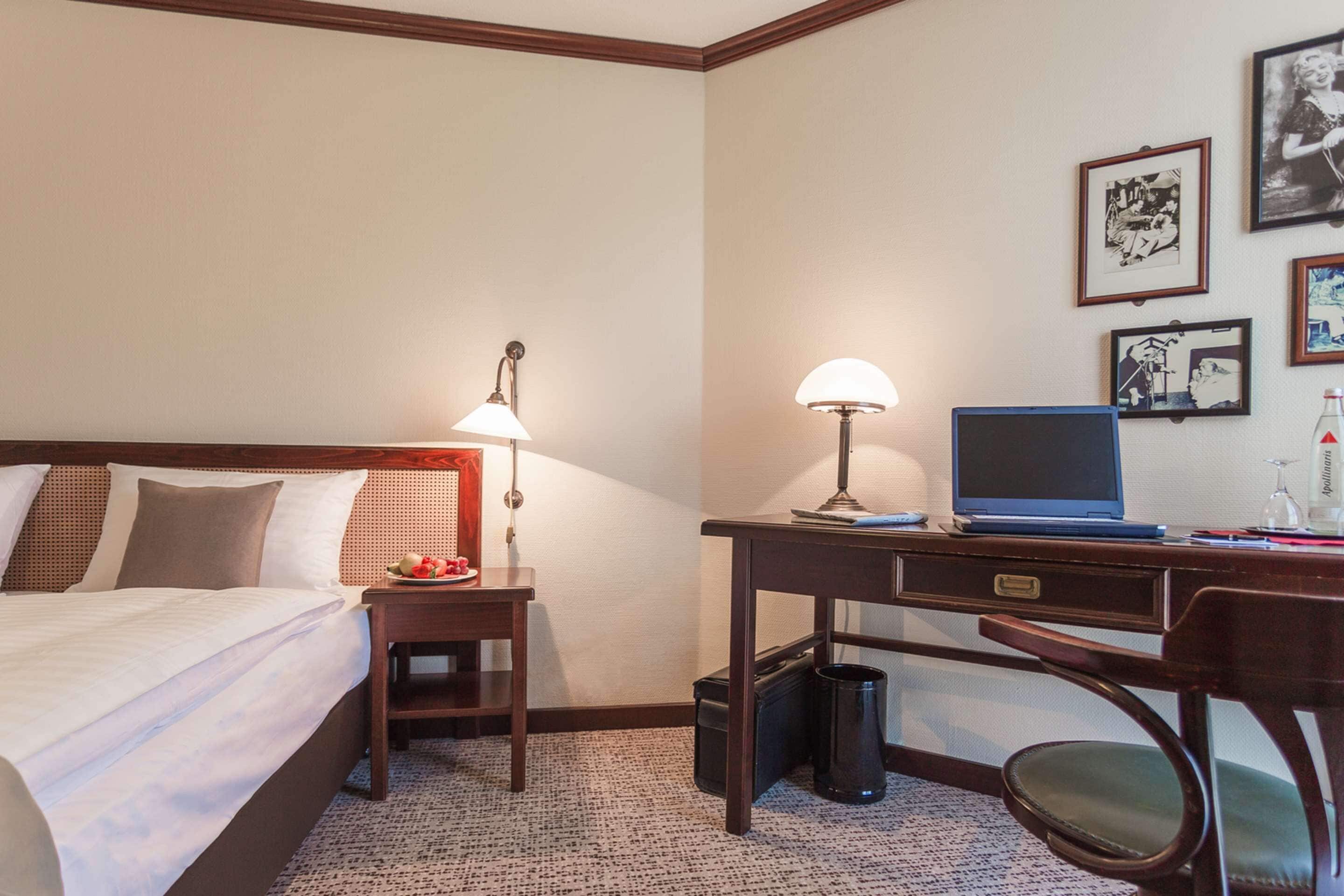 A hotel room with a bed, desk, chair, lamp, laptop, and framed pictures on the wall.