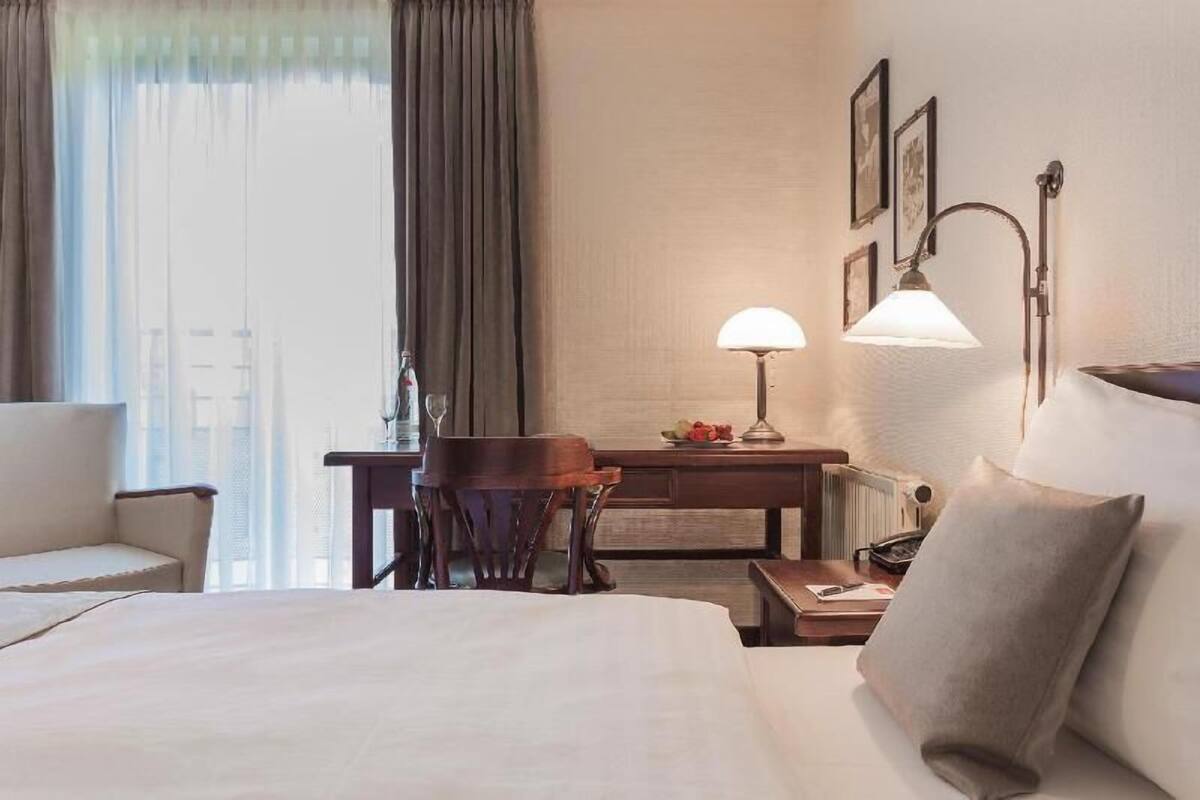 A hotel room with a bed, a desk, a chair, a lamp, and a view of the city through the window.
