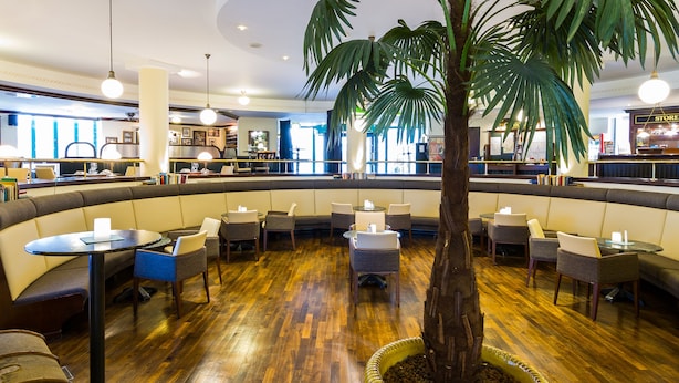 A restaurant with a central palm tree, wooden flooring, and a mix of seating options including booths and tables. - MAXX Hotel Jena (Jena)