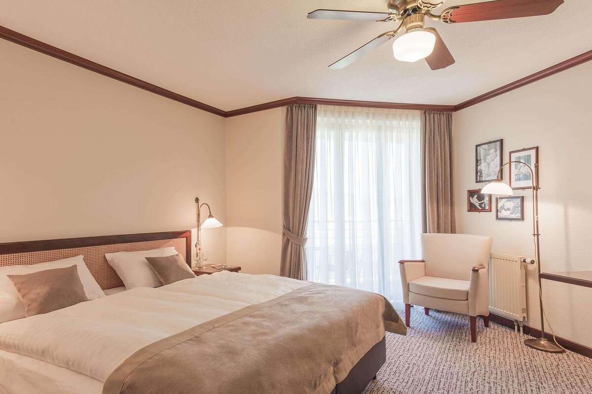 A hotel room with a large bed, a chair, a desk, and a ceiling fan.