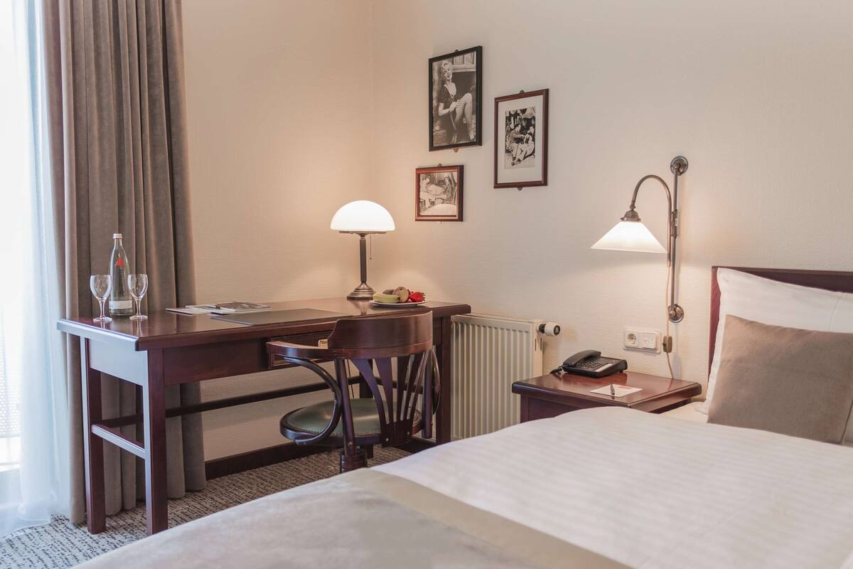 A hotel room with a bed, desk, chair, lamp, and framed pictures on the wall.