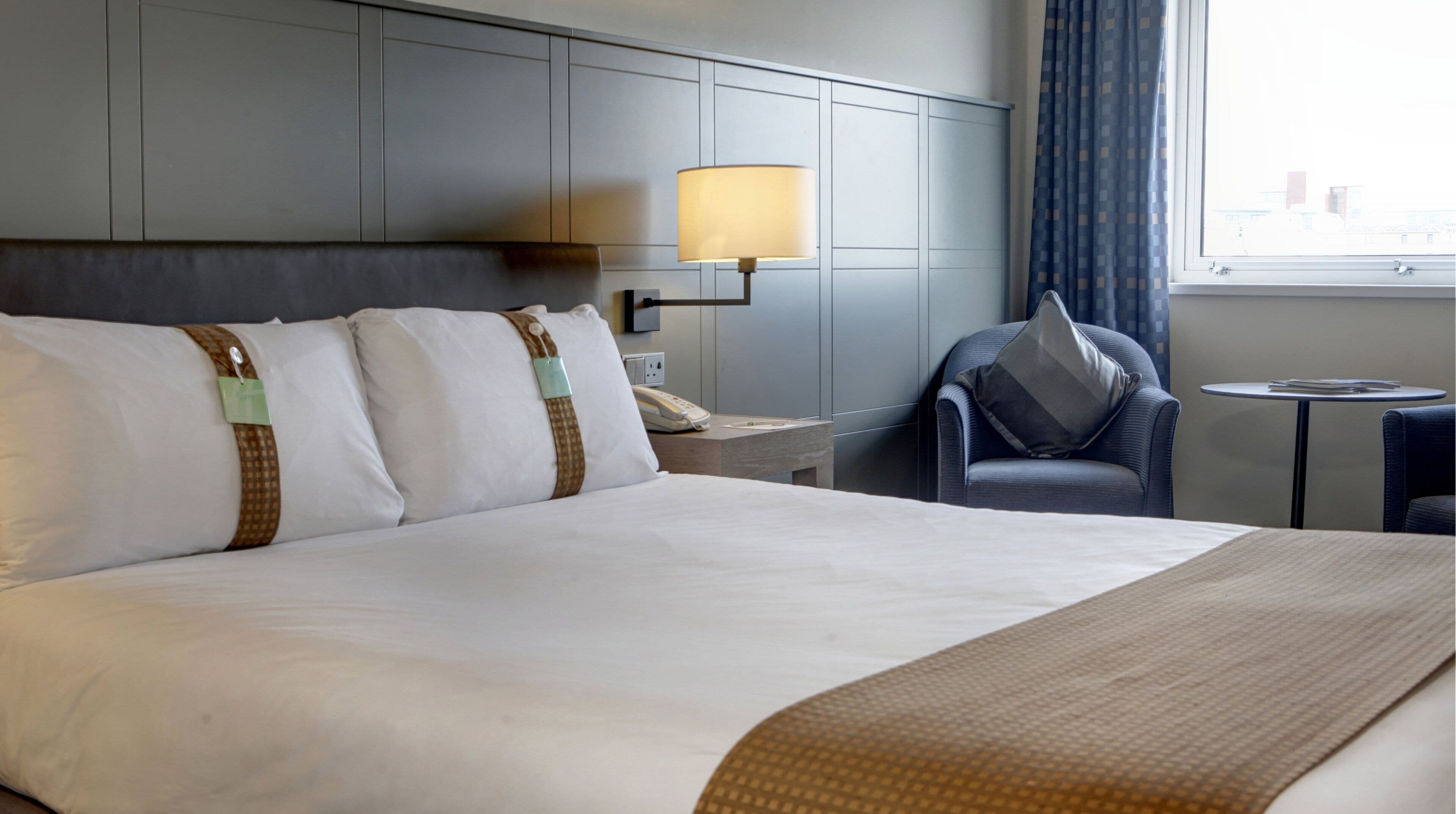 Una habitación de hotel con una cama grande, una mesita de noche con lámpara, una silla y una pequeña mesa.