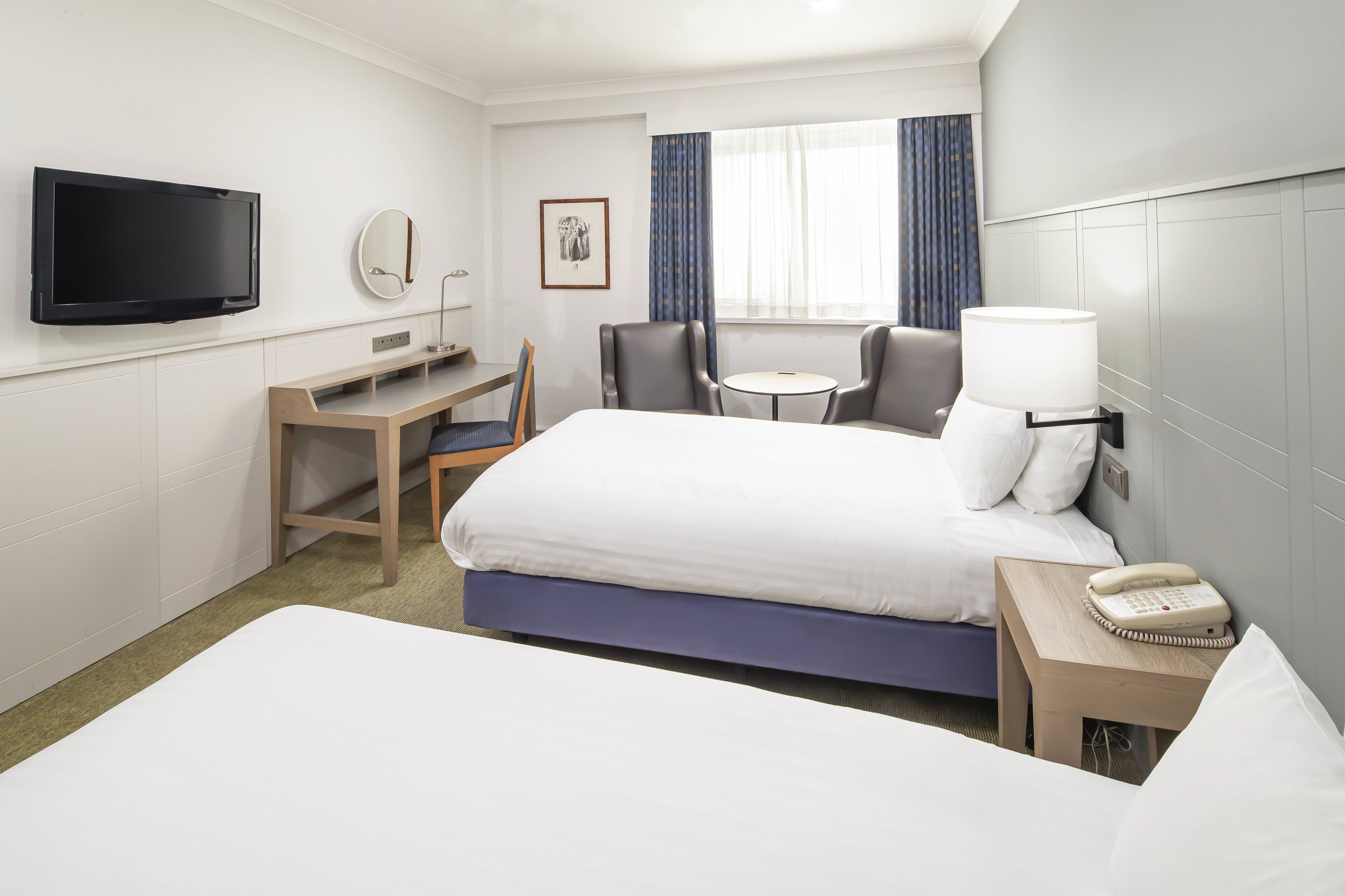 Habitación de hotel con cama, escritorio, silla, televisor, teléfono y ventana con cortinas.