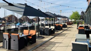 Un área de estar al aire libre con mesas y sillas, luces colgantes y vista a barcos amarrados en un puerto deportivo.