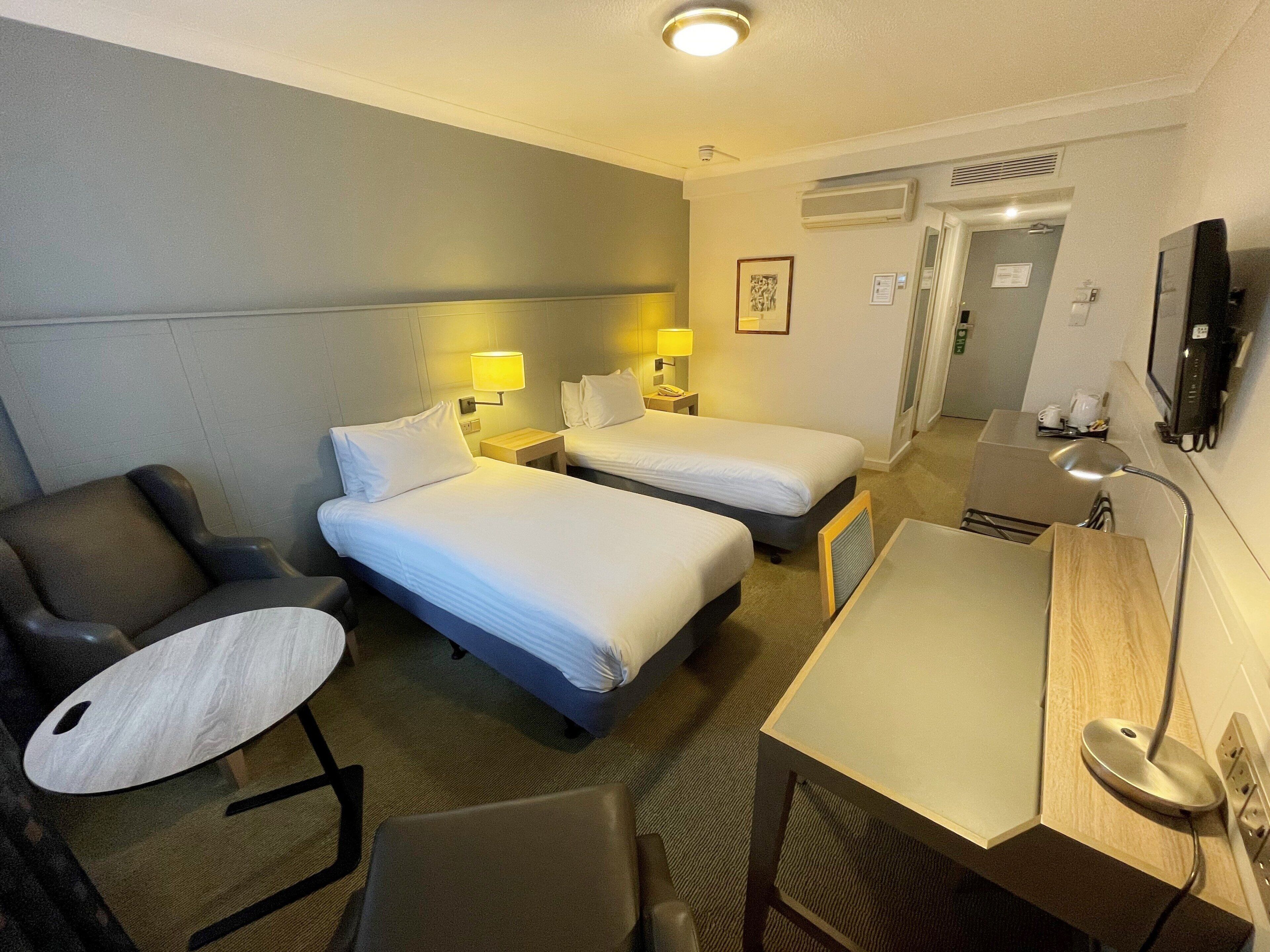 A hotel room with two single beds, a desk, a chair, a TV, and a small table.