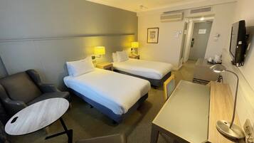 A hotel room with two single beds, a desk, a chair, a TV, and a small table.