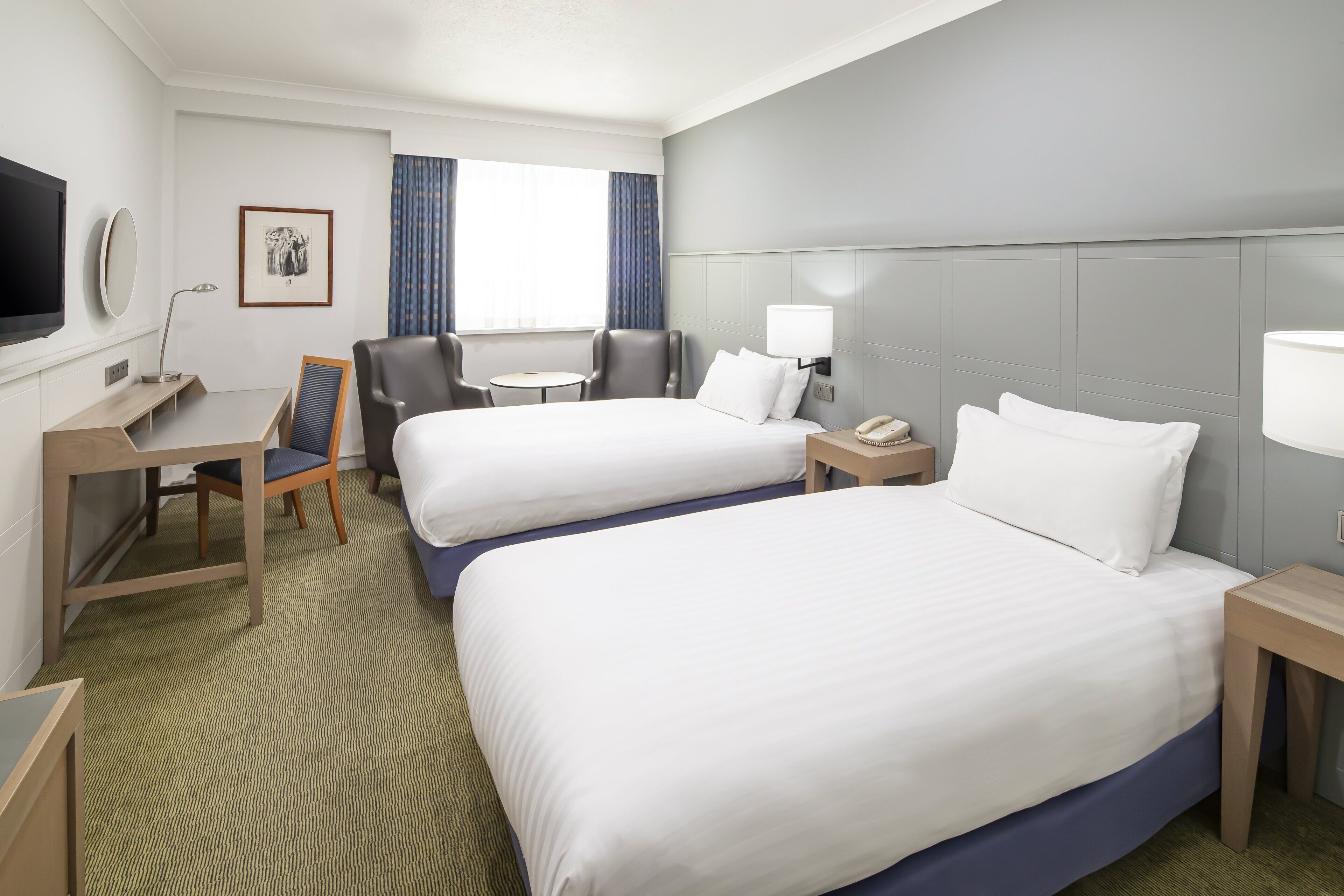 A hotel room with two beds, a desk, a chair, a TV, and a window with curtains.