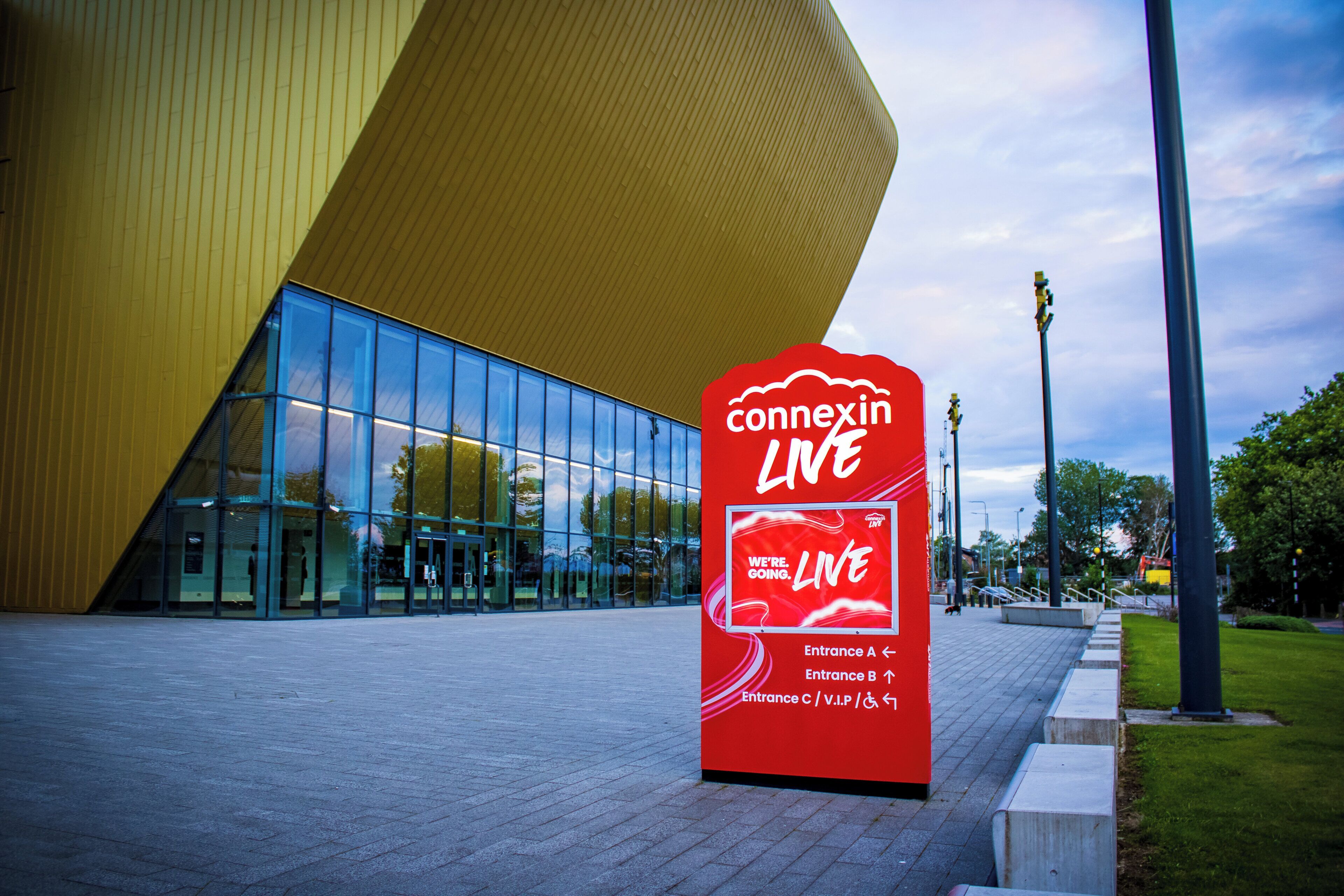Edifício moderno com fachada de vidro ampla e placa vermelha indicando 'connexin LIVE' com setas direcionais para as entradas.