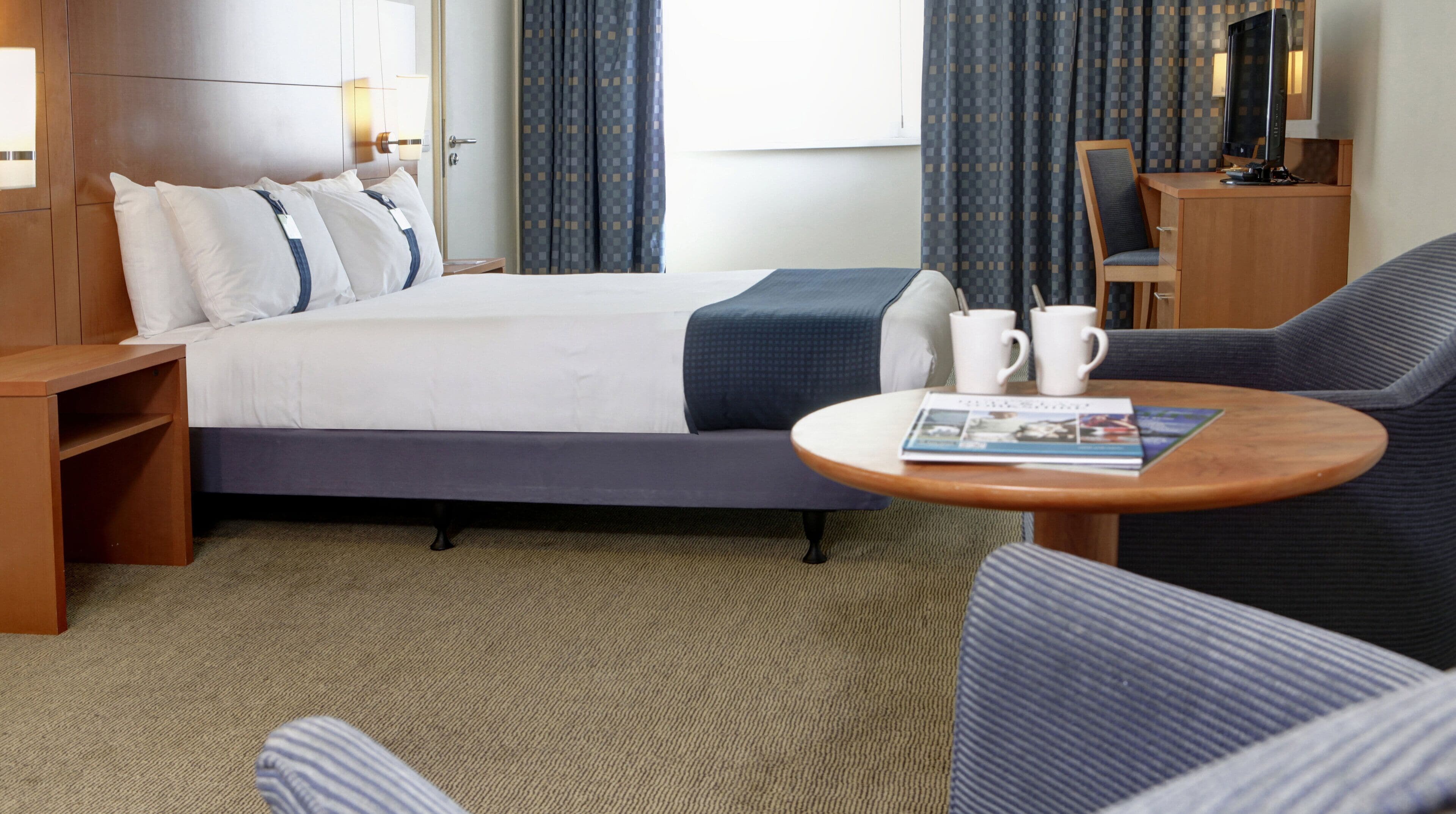 A hotel room with a bed, a sofa, a small table with a magazine, a TV, and a desk with a computer.