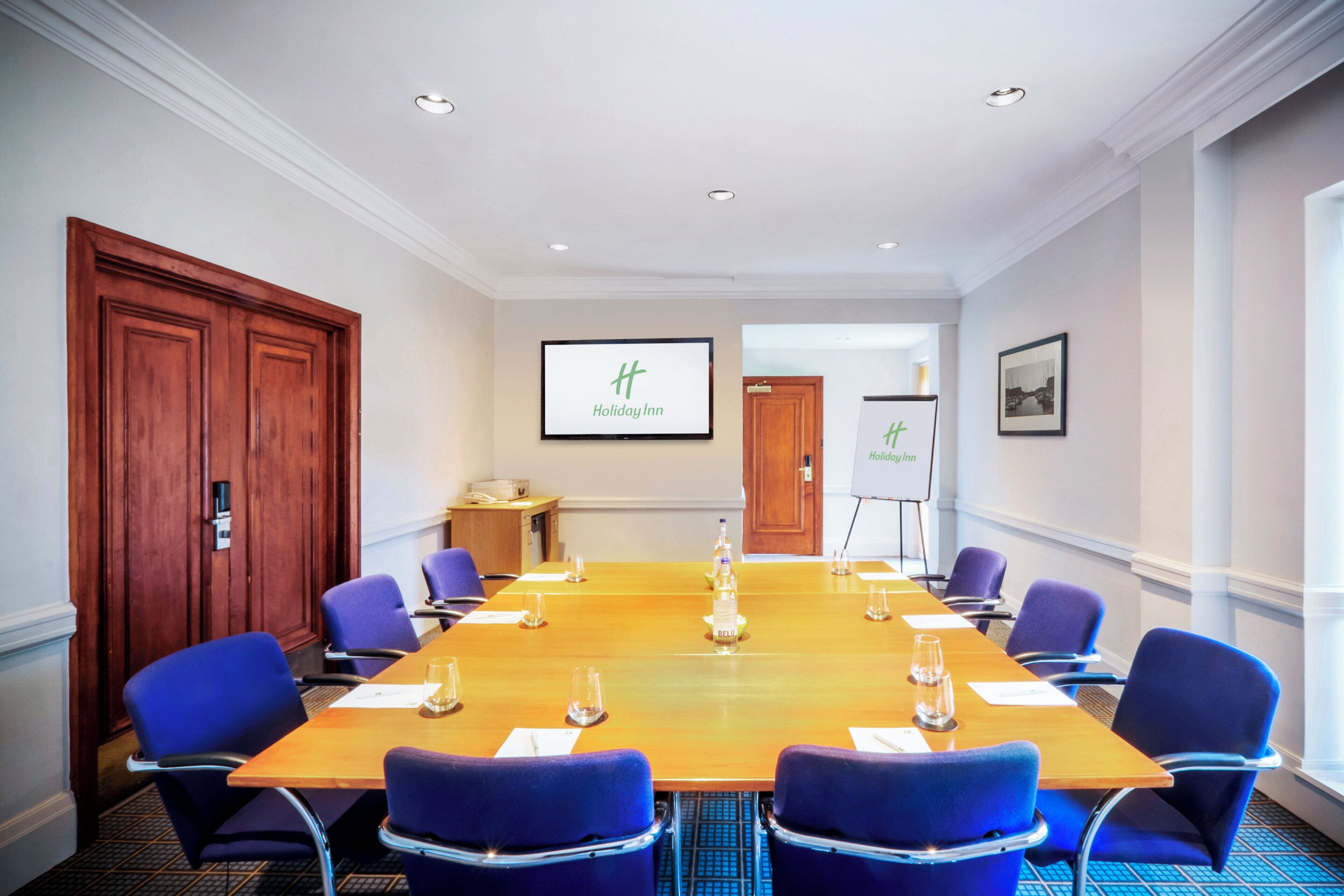 Une salle de conférence avec une longue table, des chaises bleues, une porte en bois et un tableau blanc sur lequel est écrit « Holiday Inn ».