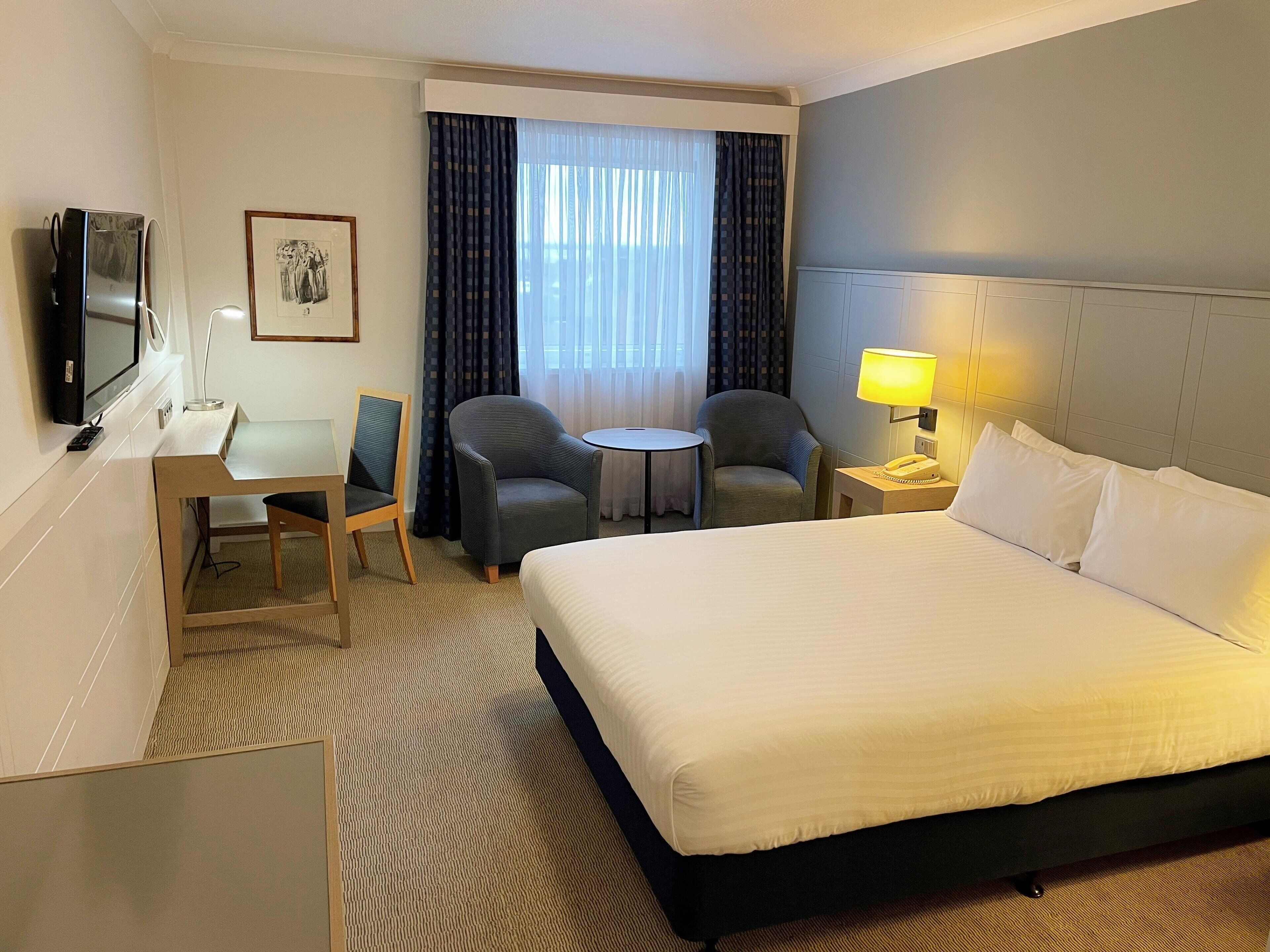 A hotel room with a large bed, a desk with a chair, a small table, a TV, and a lamp.
