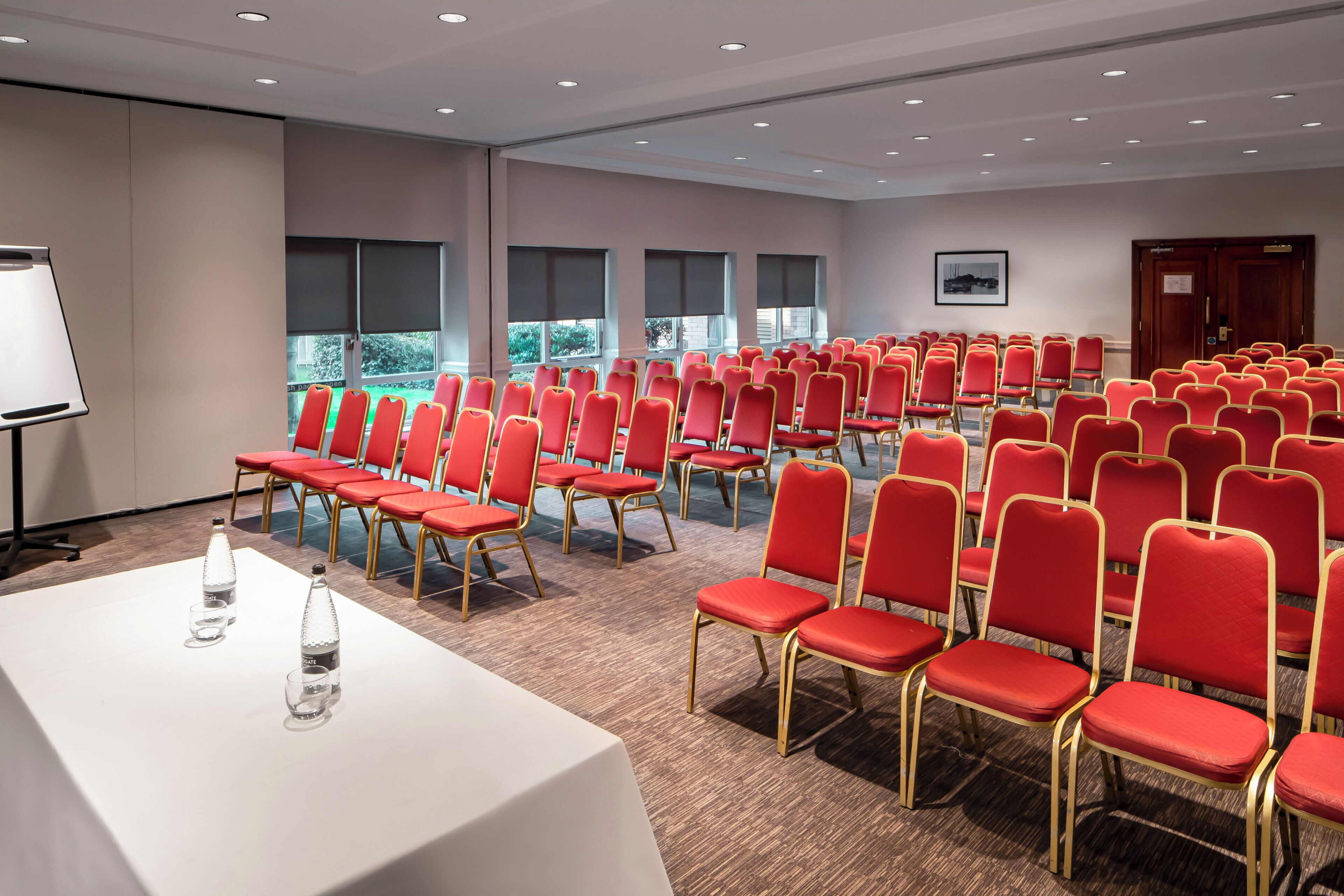 A conference room with rows of red chairs, a white table with water bottles, and a whiteboard.
