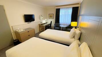 A hotel room with two beds, a desk, a chair, a TV, a lamp, and a window with curtains.