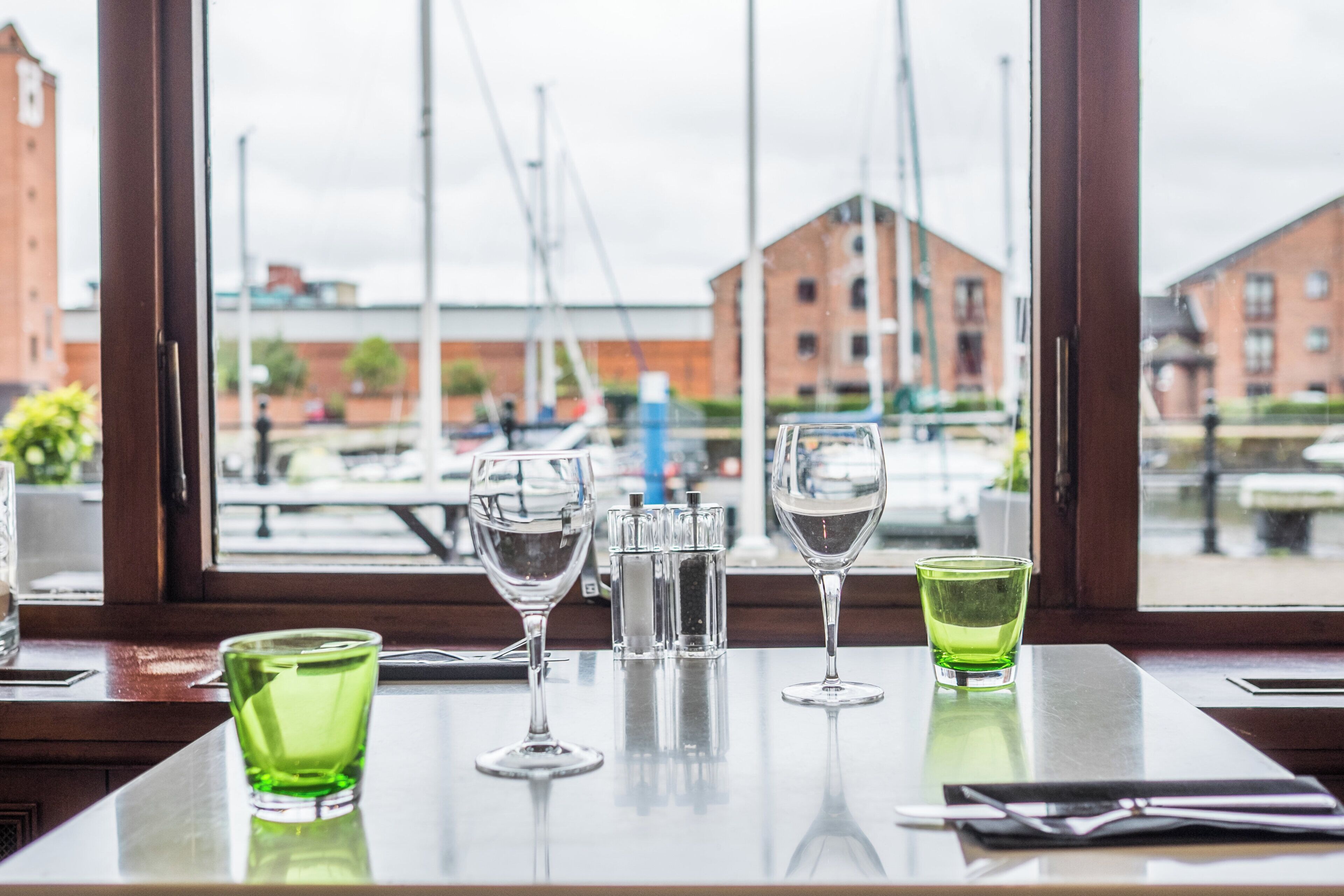 Una mesa con vasos y una botella, una ventana con vistas a edificios y barcos, y una mesa de madera.