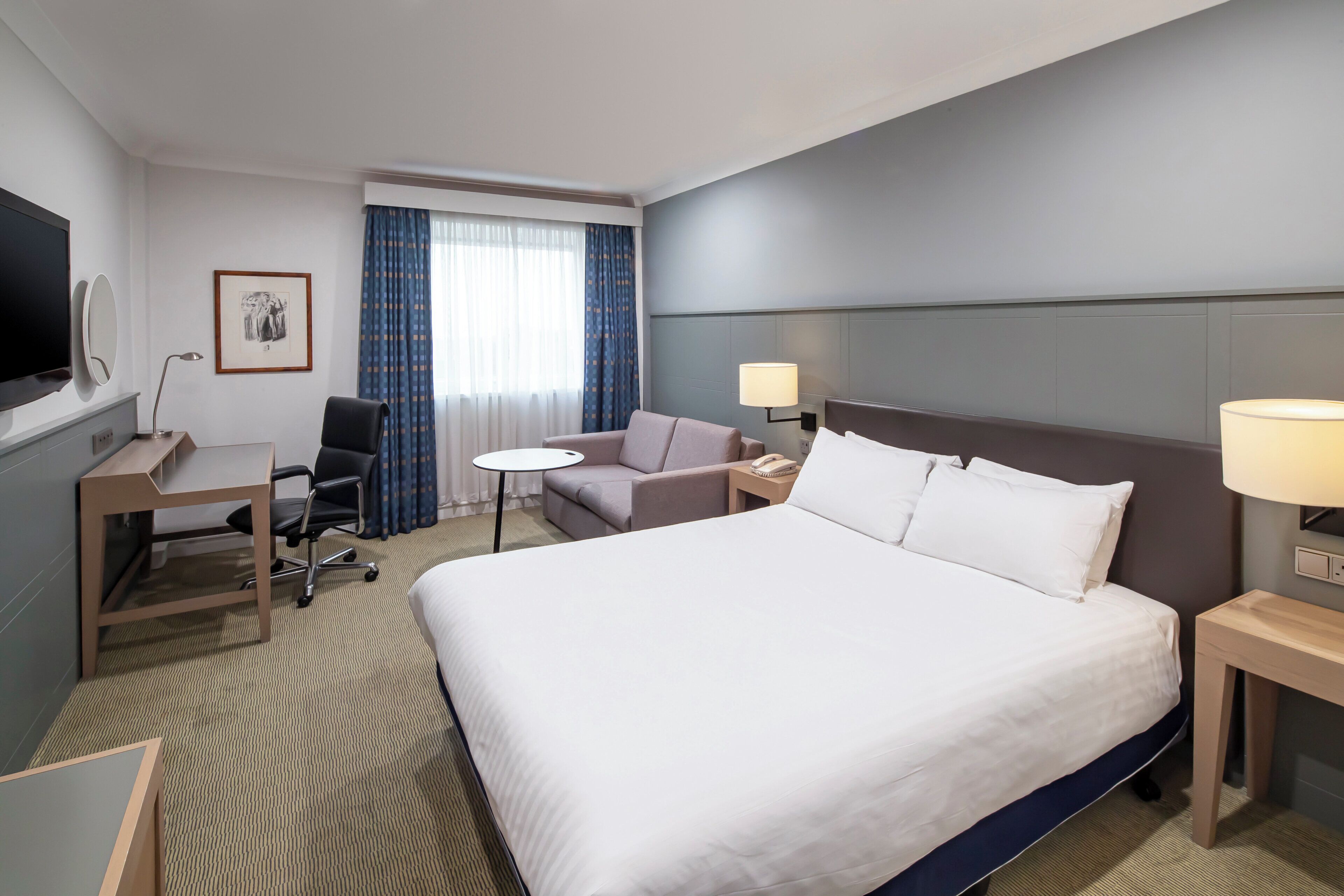 A hotel room with a large bed, a desk, a chair, a small table, a lamp, and a television.