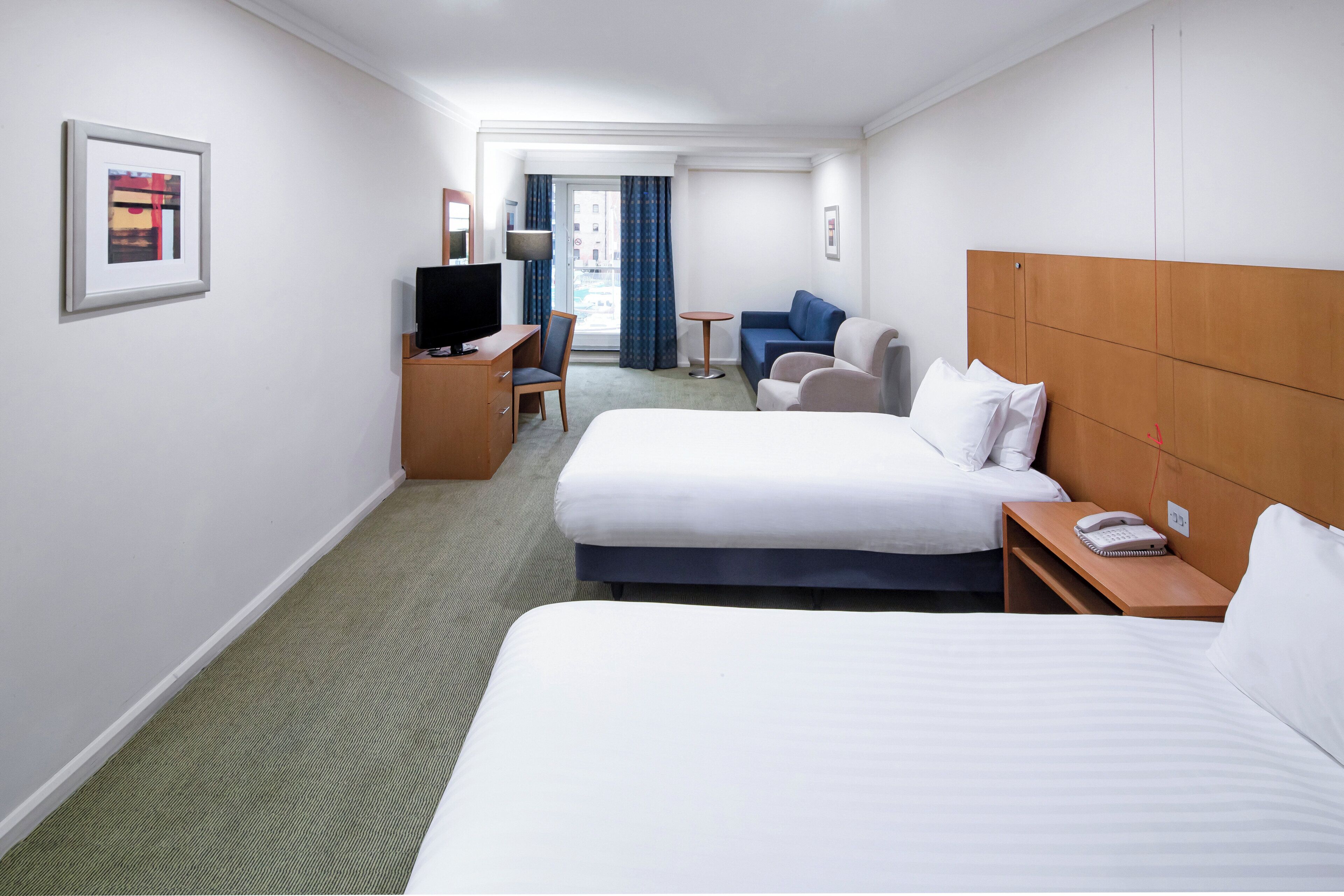 Habitación de hotel con dos camas, un escritorio con televisor, una silla, una mesita y una ventana con cortinas.