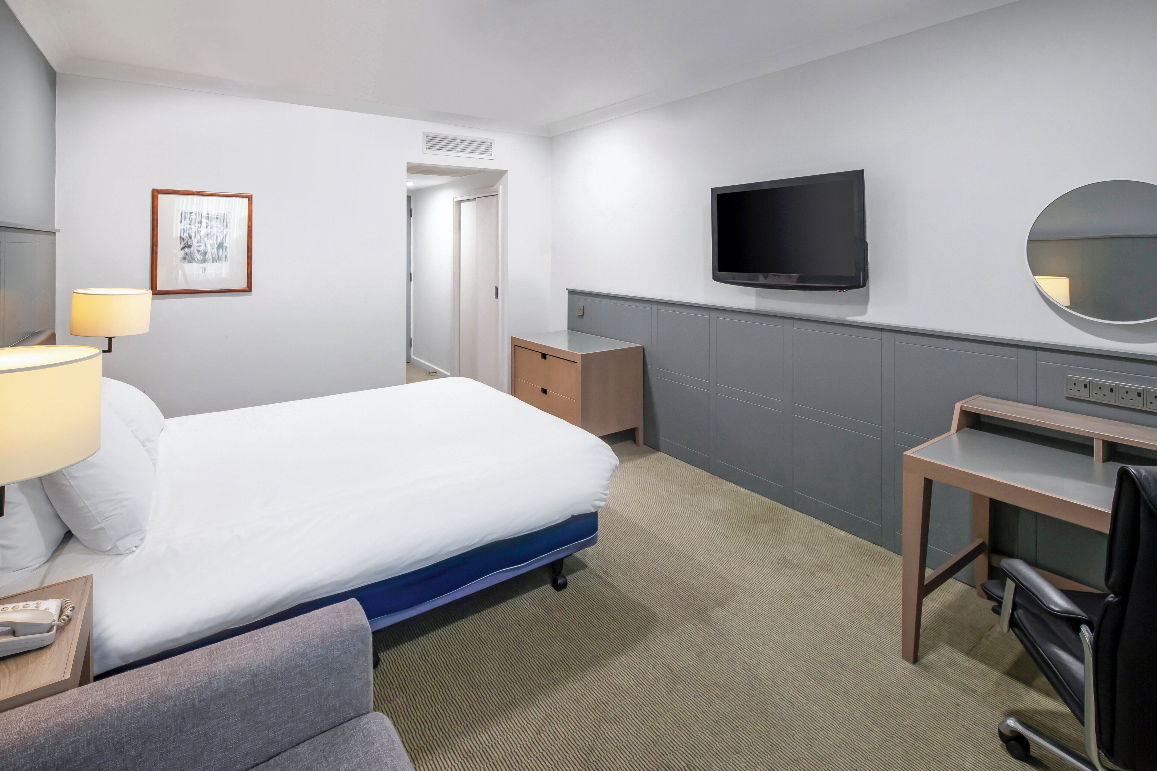 Una habitación de hotel con cama, mesita de noche, lámpara, escritorio, silla, televisor y espejo.
