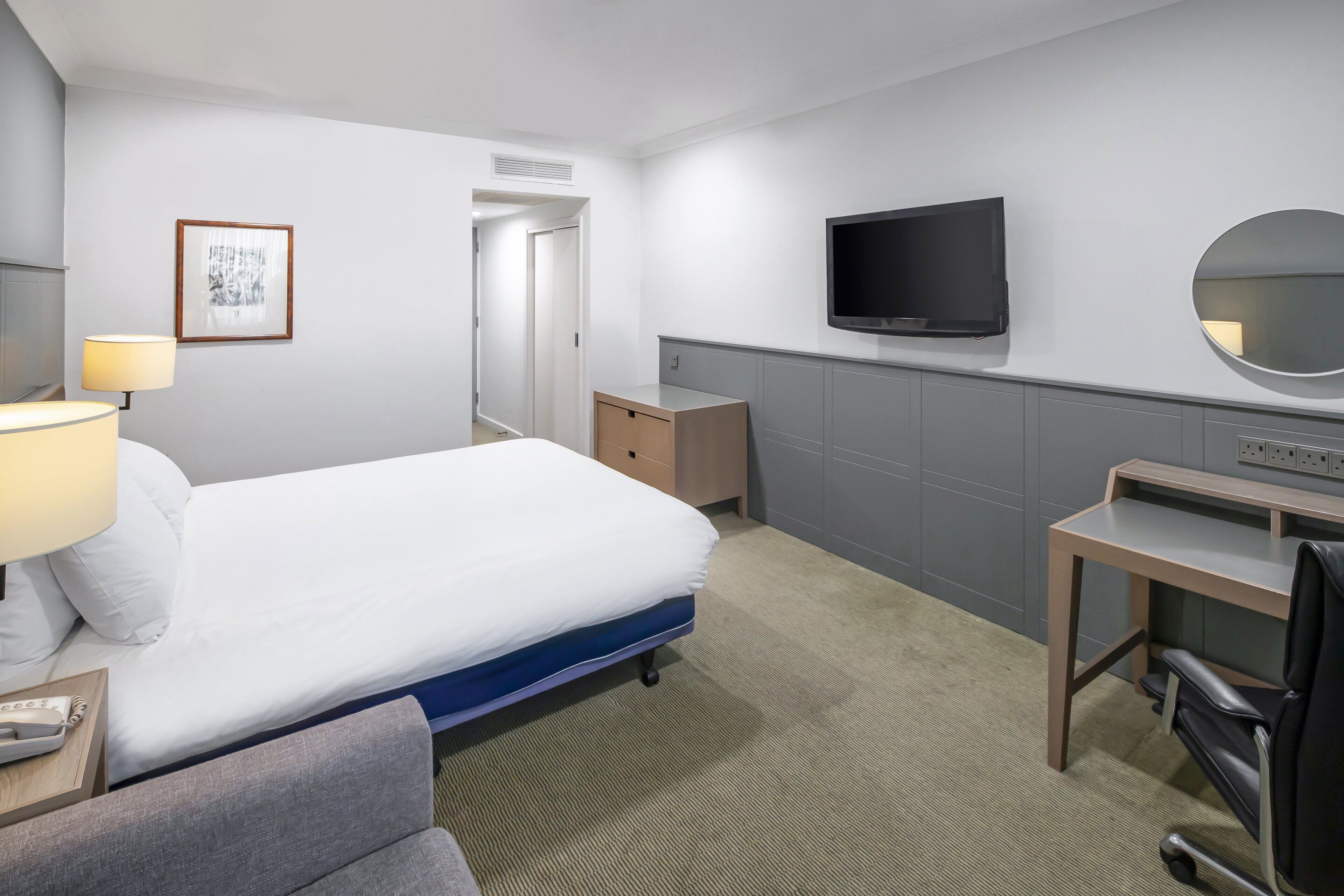 A hotel room with a bed, a desk, a chair, a TV, a mirror, a lamp, and a telephone.