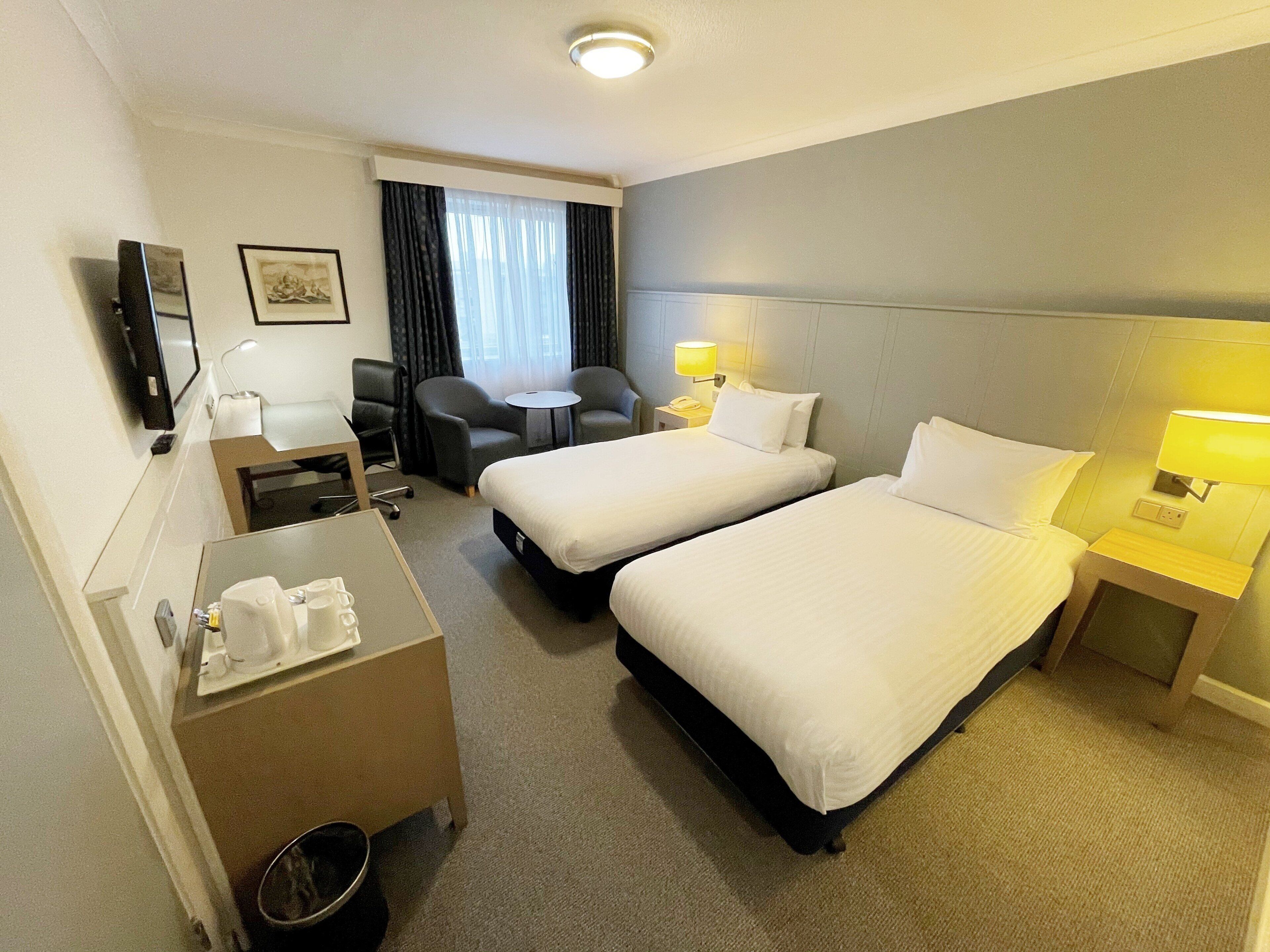 A hotel room with two beds, a desk, a chair, a small table, a TV, and a window with curtains.