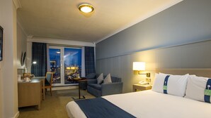 Una habitación de hotel con una cama grande, un escritorio, una silla, un sofá y vistas a la ciudad de noche.