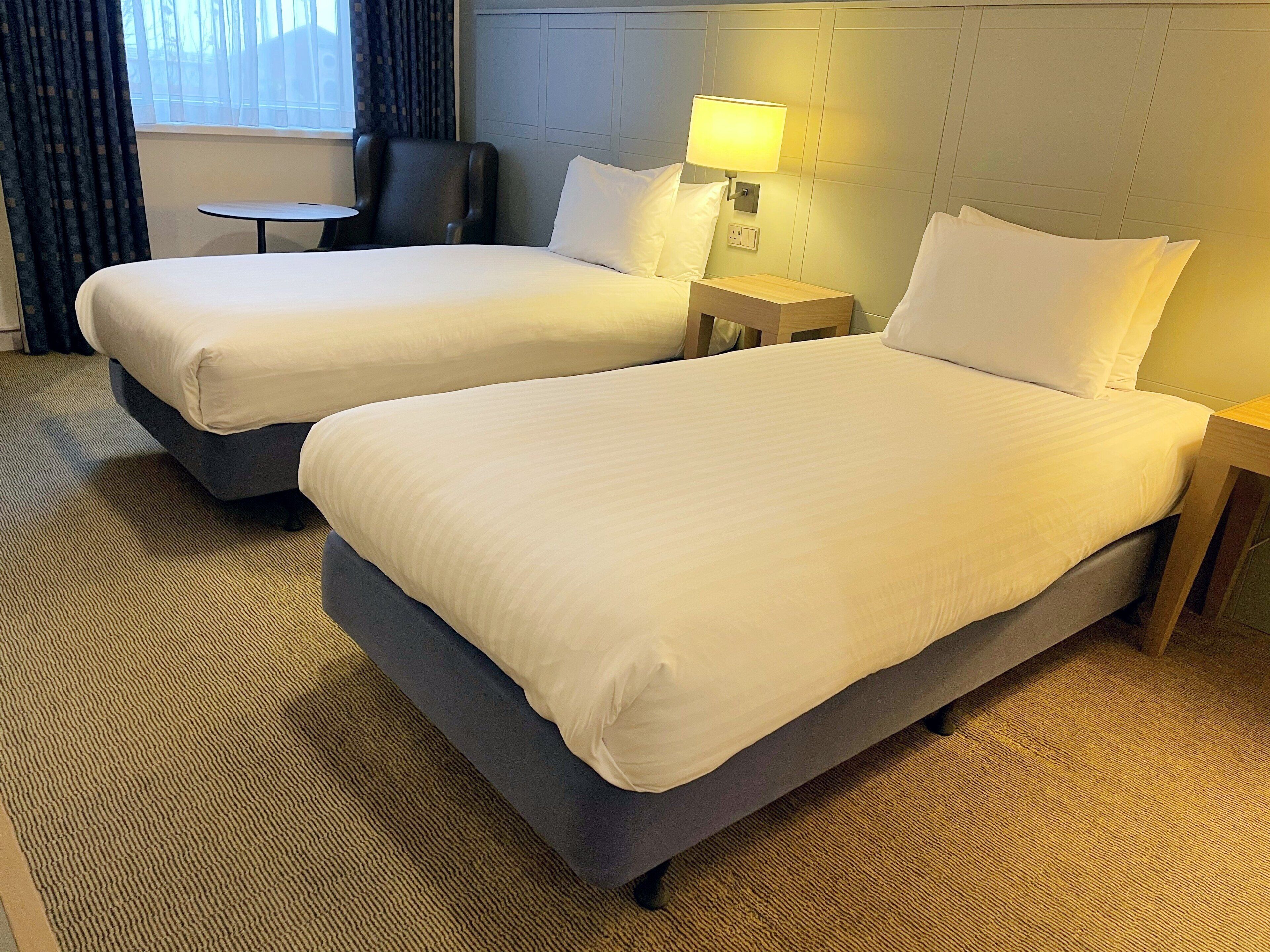 A hotel room with two beds, a small table, a chair, and a lamp.