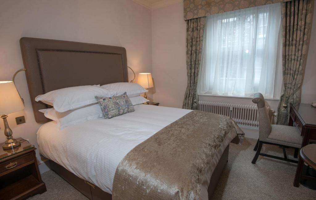 Een keurig opgemaakt bed met witte lakens en een bruine plaid, een houten nachtkastje met een lamp, een stoel en een raam met dunne gordijnen.