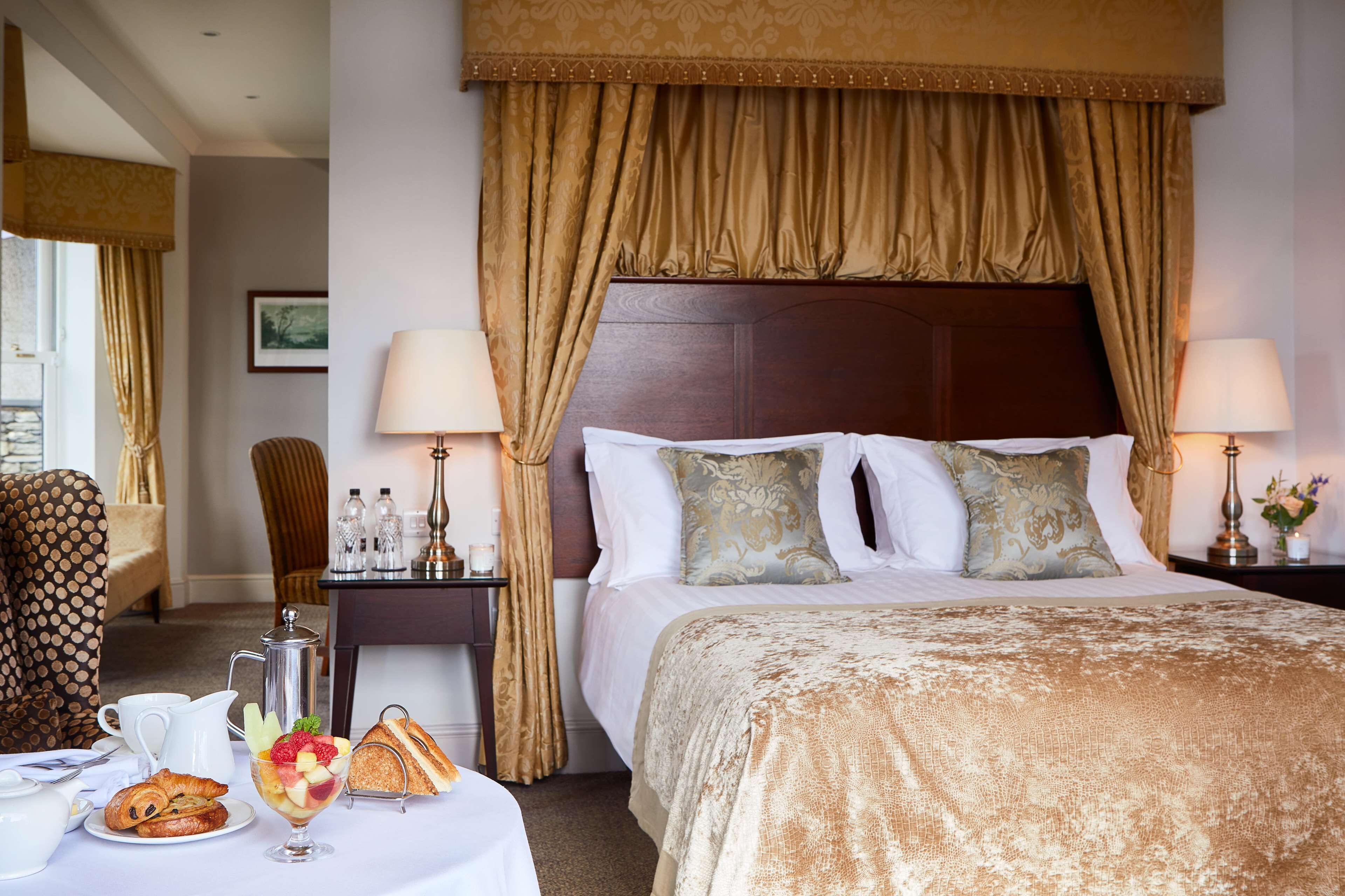 A hotel room with a large bed, two bedside tables with lamps, a dining table with breakfast items, and a patterned armchair.