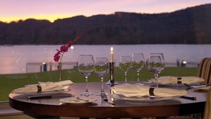 A table set for a meal with wine glasses, a candle, and a vase with flowers, overlooking a body of water and hills. - Macdonald Old England Hotel & Spa (Windermere)