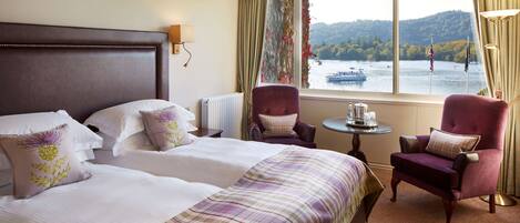 A hotel room with a large bed, two armchairs, a small table, and a view of a lake with boats.