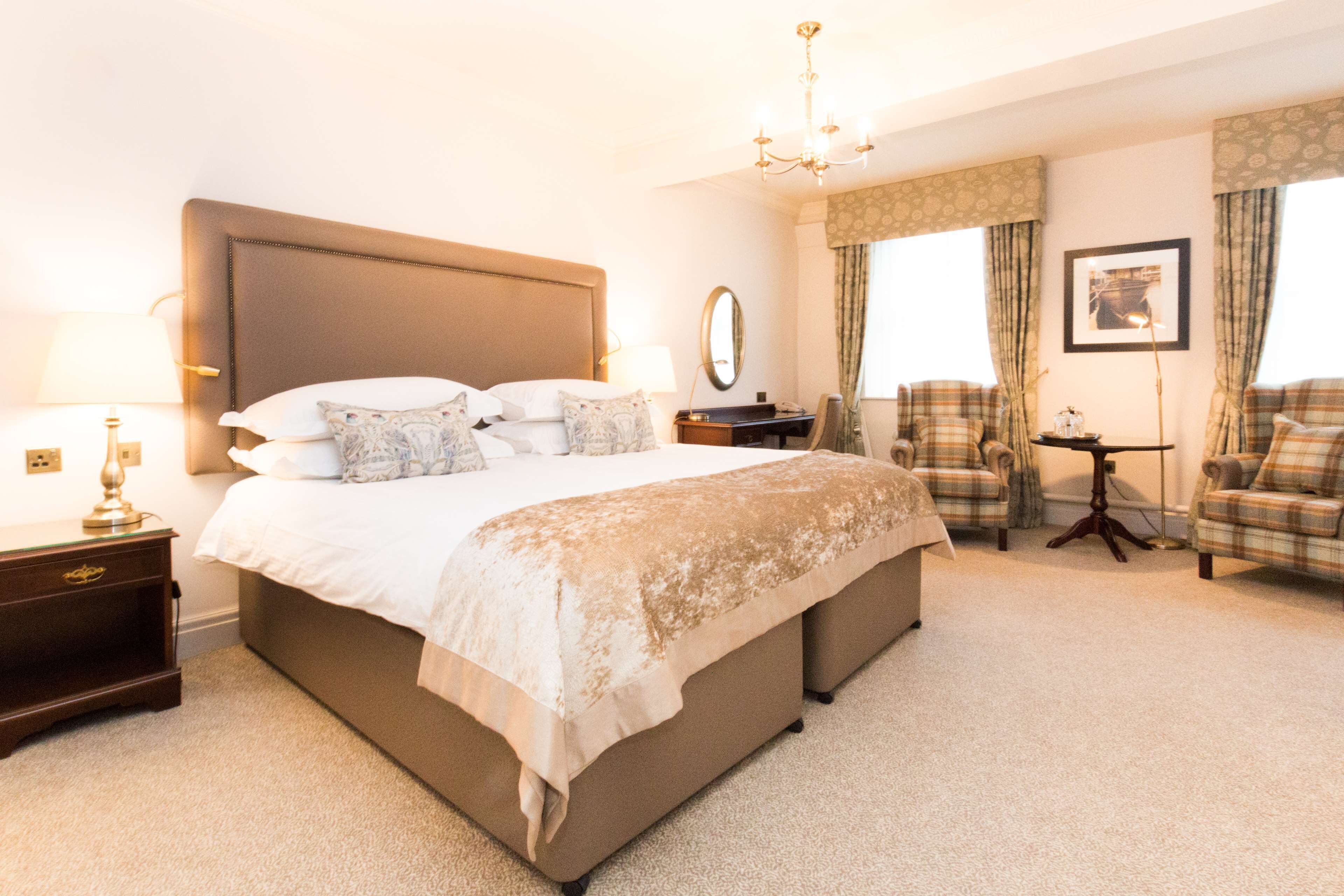 A hotel room with a large bed, a nightstand, a chair, a desk, and a chandelier.