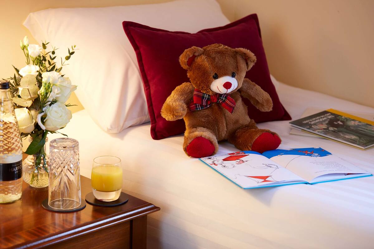 A bed with a red pillow, a teddy bear, a book, a glass of orange juice, and a vase with white flowers.